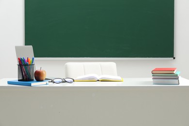 Photo of Different school stationery, glasses and apple on white table near blackboard indoors