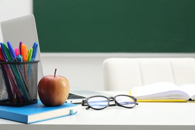 Photo of Different school stationery, laptop, glasses and apple on white table near blackboard indoors, closeup