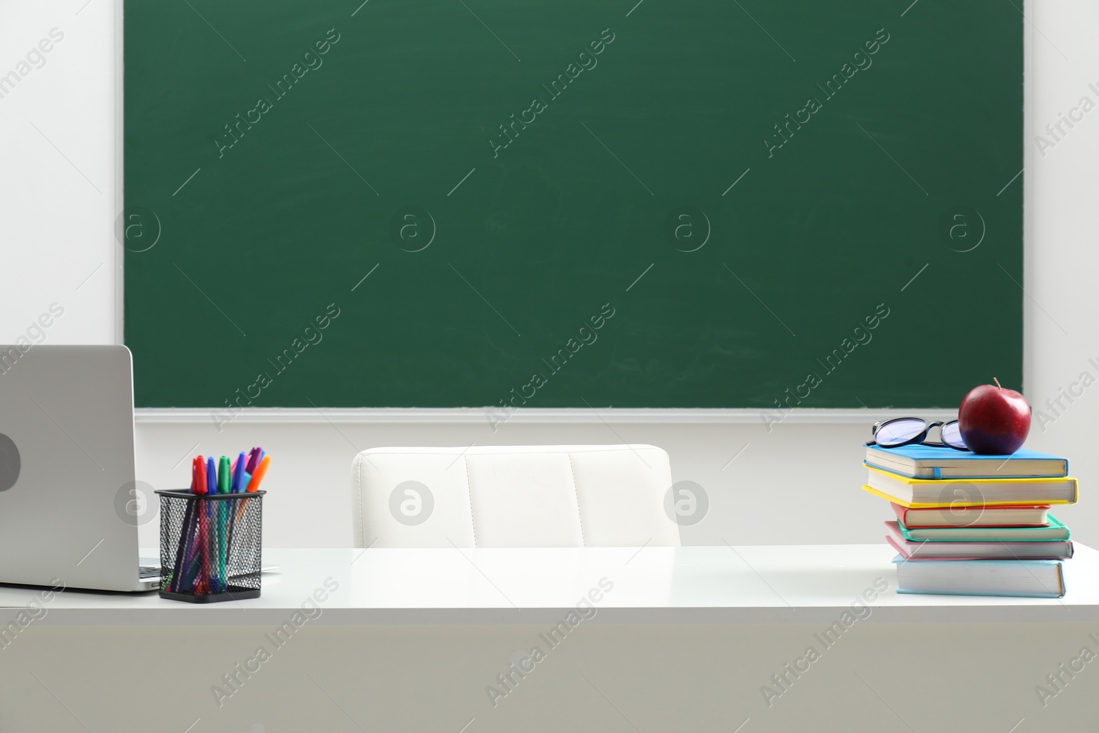 Different school stationery, laptop, glasses and red apple on white table near blackboard indoors Photo of Different school stationery, laptop, glasses and red apple on white table near blackboard indoors