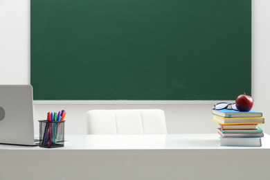 Photo of Different school stationery, laptop, glasses and red apple on white table near blackboard indoors