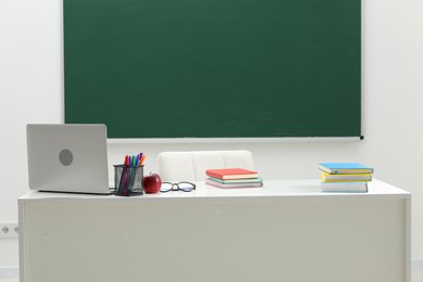 Photo of Different school stationery, laptop, glasses and red apple on white table near blackboard indoors