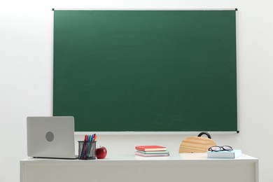Photo of Different school stationery, laptop, glasses and red apple on white table near blackboard indoors