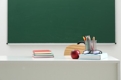 Photo of Different school stationery, glasses and red apple on white table near blackboard indoors