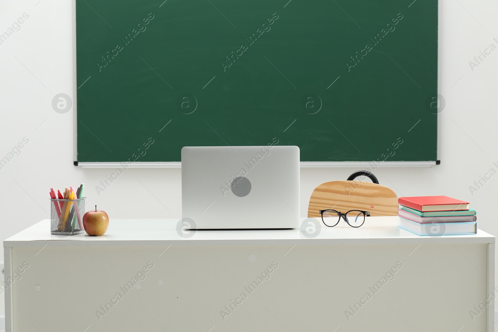 Different school stationery, laptop, glasses and apple on white table near blackboard indoors Photo of Different school stationery, laptop, glasses and apple on white table near blackboard indoors
