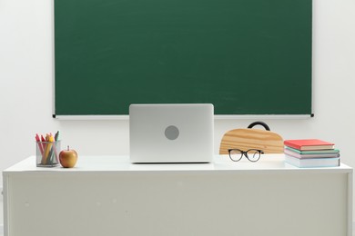 Photo of Different school stationery, laptop, glasses and apple on white table near blackboard indoors
