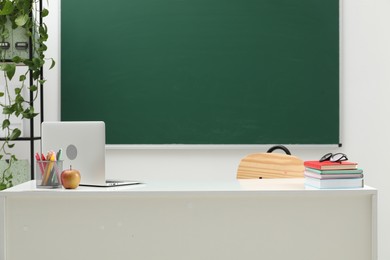 Photo of Different school stationery, laptop, glasses and apple on white table near blackboard indoors