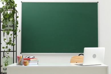 Photo of Different school stationery, laptop, glasses and apple on white table near blackboard indoors