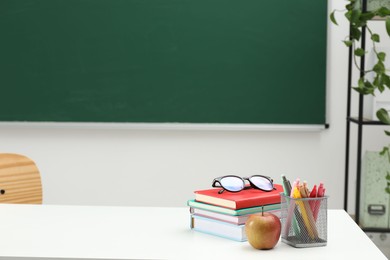 Photo of Different school stationery, glasses and apple on white table near blackboard indoors, space for text