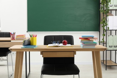 Photo of Different school stationery, glasses and red apple on wooden table near blackboard indoors