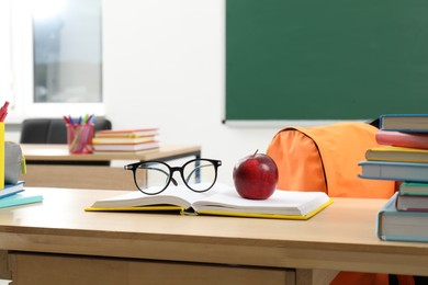 Photo of Different school stationery, glasses and red apple on wooden table near blackboard indoors