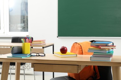 Photo of Different school stationery, glasses and red apple on wooden table near blackboard indoors