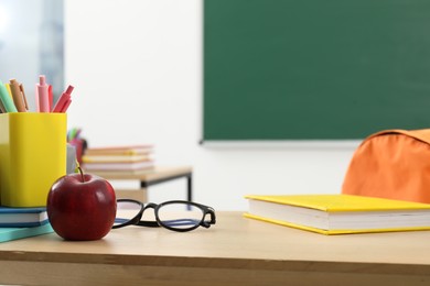 Photo of Different school stationery, glasses and red apple on wooden table near blackboard indoors