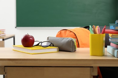 Photo of Different school stationery, glasses and red apple on wooden table near blackboard indoors