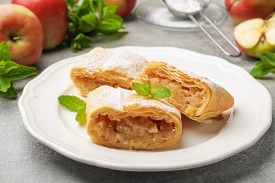 Photo of Pieces of tasty apple strudel with powdered sugar, mint and fruits on grey table, closeup