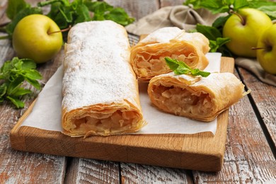 Photo of Tasty apple strudels with powdered sugar, mint and fruits on wooden table, closeup