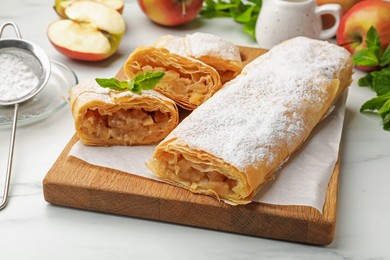 Photo of Tasty apple strudels with powdered sugar, mint and fruits on white marble table, closeup
