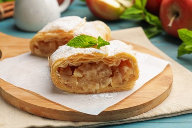 Photo of Pieces of tasty apple strudel with powdered sugar, mint and fruits on light blue wooden table, closeup