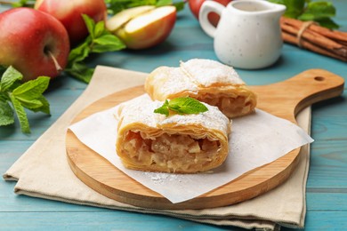 Photo of Pieces of tasty apple strudel with powdered sugar, mint, fruits and cinnamon on light blue wooden table, closeup