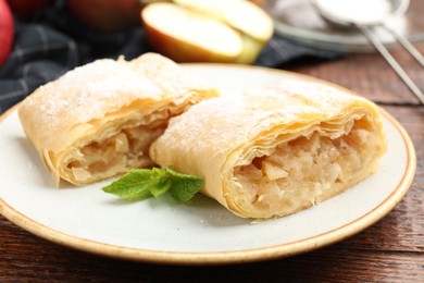 Photo of Pieces of tasty apple strudel with powdered sugar, mint and fruit on wooden table, closeup