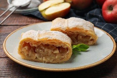 Photo of Pieces of tasty apple strudel with powdered sugar, mint and fruits on wooden table, closeup