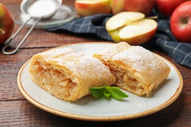 Photo of Pieces of tasty apple strudel with powdered sugar, mint and fruits on wooden table, closeup