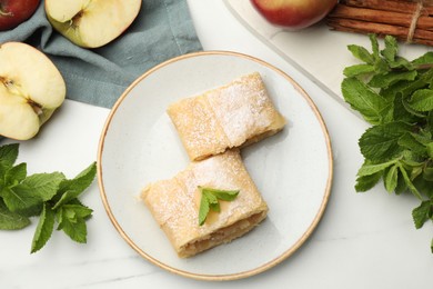 Photo of Pieces of tasty apple strudel with powdered sugar, mint, fruits and cinnamon on white marble table, flat lay