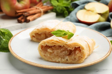 Photo of Pieces of tasty apple strudel with powdered sugar, mint, fruits and cinnamon on white marble table, closeup
