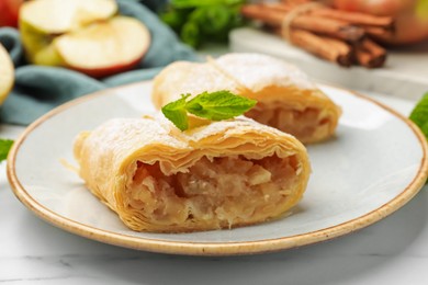 Photo of Pieces of tasty apple strudel with powdered sugar, mint, fruit and cinnamon on white marble table, closeup