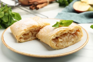 Photo of Pieces of tasty apple strudel with powdered sugar, mint, fruit and cinnamon on white marble table, closeup