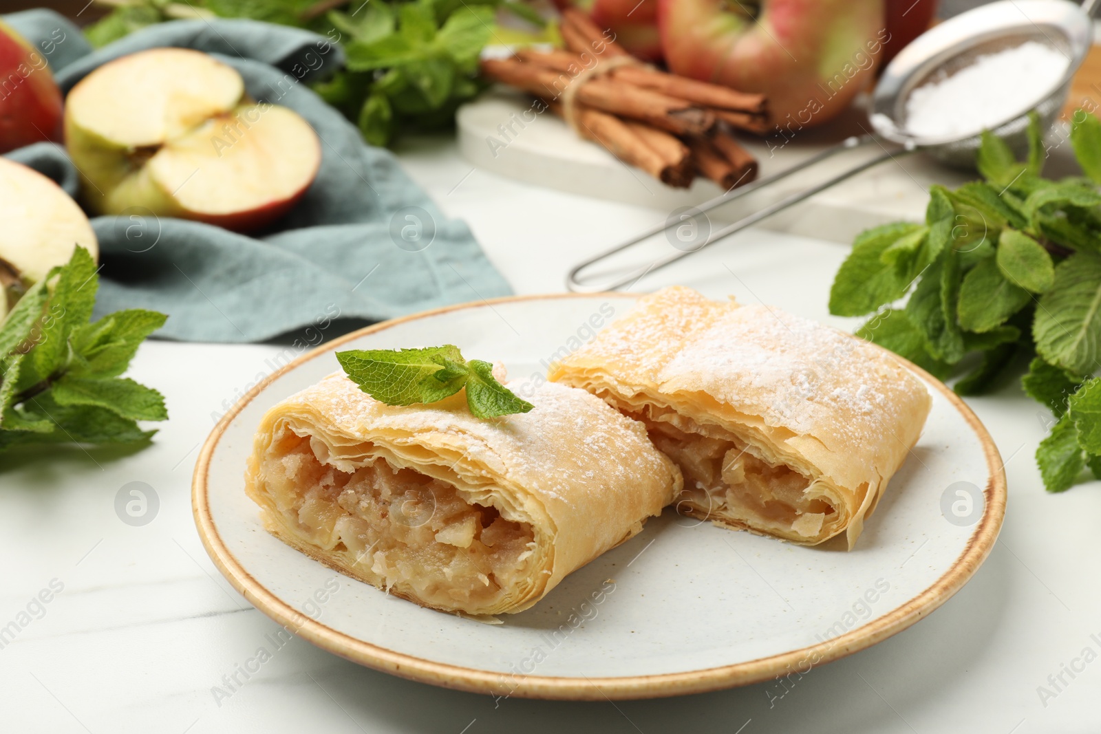 Pieces of tasty apple strudel with powdered sugar, mint, fruits and cinnamon on white marble table, closeup Photo of Pieces of tasty apple strudel with powdered sugar, mint, fruits and cinnamon on white marble table, closeup