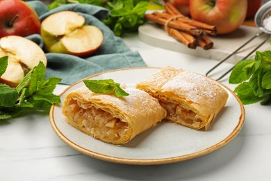 Photo of Pieces of tasty apple strudel with powdered sugar, mint, fruits and cinnamon on white marble table, closeup