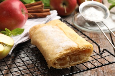 Photo of Tasty apple strudel with powdered sugar, mint, fruits and cinnamon on wooden table, closeup