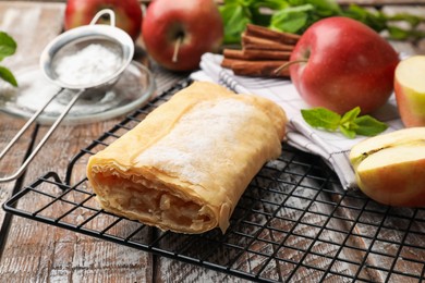 Photo of Tasty apple strudel with powdered sugar, mint, fruits and cinnamon on wooden table, closeup