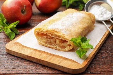 Photo of Tasty apple strudel with powdered sugar, mint and fruits on wooden table, closeup