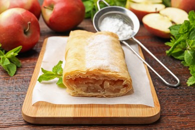 Photo of Tasty apple strudel with powdered sugar, mint and fruits on wooden table, closeup