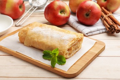 Photo of Tasty apple strudel with powdered sugar, mint, fruits and cinnamon on wooden table, closeup