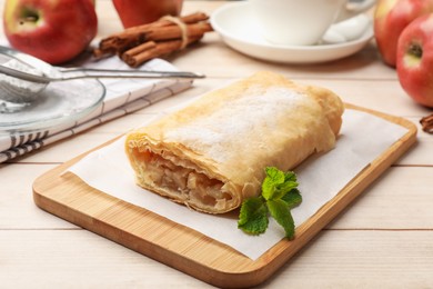 Photo of Tasty apple strudel with powdered sugar, mint, fruits and cinnamon on wooden table, closeup