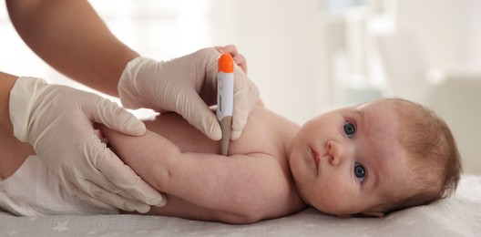 Photo of Pediatrician examining baby with thermometer at table in clinic, closeup