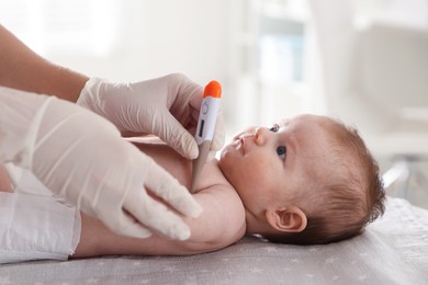 Photo of Pediatrician examining baby with thermometer at table in clinic, closeup