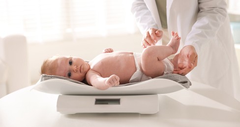 Photo of Pediatrician weighting baby with scales at white table in clinic, closeup