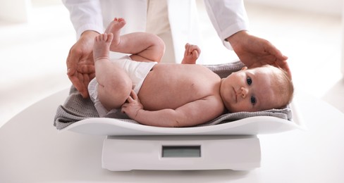 Photo of Pediatrician weighting baby with scales at white table in clinic, closeup