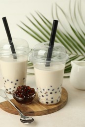 Photo of Tasty milk bubble tea in plastic cups, tapioca pearls and green leaves on beige table, closeup