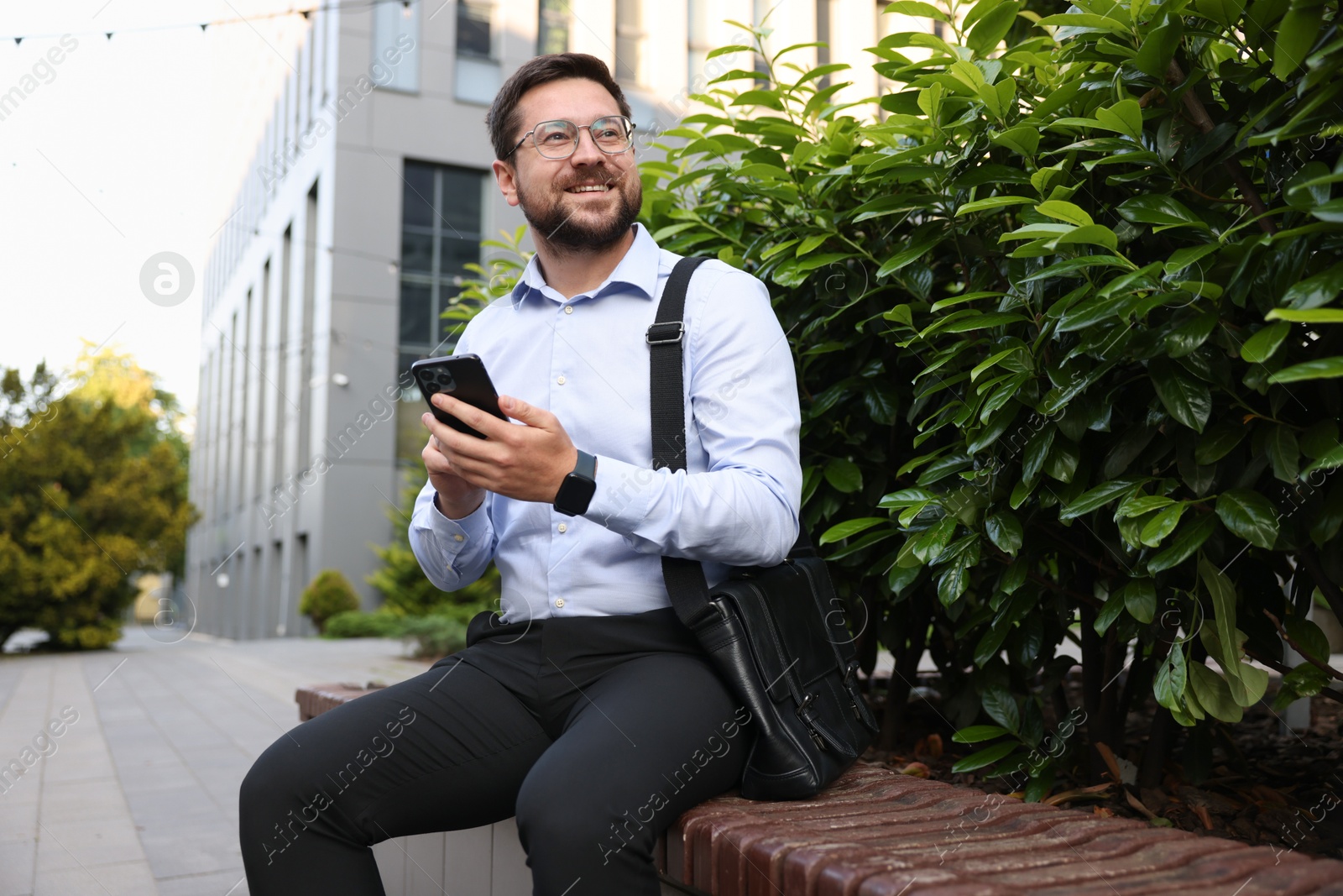 Successful businessman with smartphone on bench near office building Photo of Successful businessman with smartphone on bench near office building