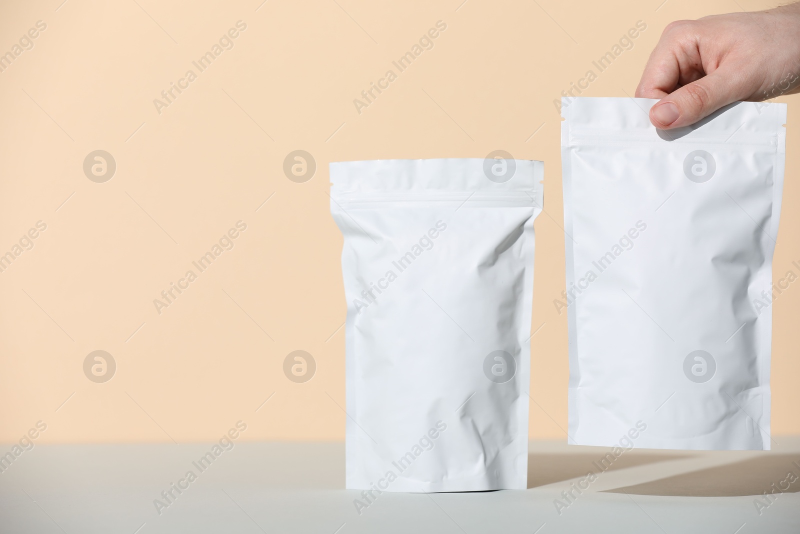 Photo of Woman holding blank zipper stand-up pouches at grey table against beige background, closeup. Space for text