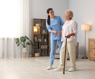 Photo of Nurse assisting senior woman with walking cane indoors, space for text. Home health care service