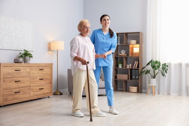 Photo of Nurse assisting senior woman with walking cane indoors. Home health care service