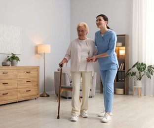 Photo of Nurse assisting senior woman with walking cane indoors. Home health care service