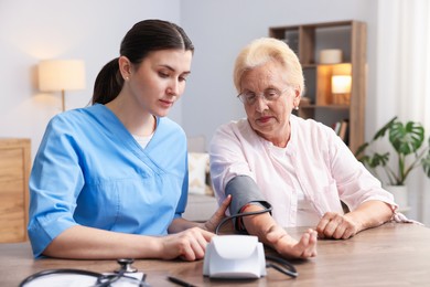 Photo of Nurse measuring patient's blood pressure at wooden table indoors. Home health care service