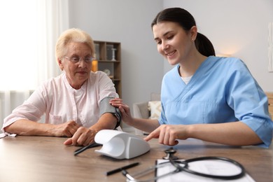 Photo of Nurse measuring patient's blood pressure at wooden table indoors. Home health care service