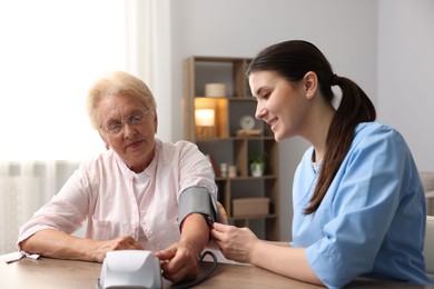Photo of Nurse measuring patient's blood pressure at wooden table indoors. Home health care service